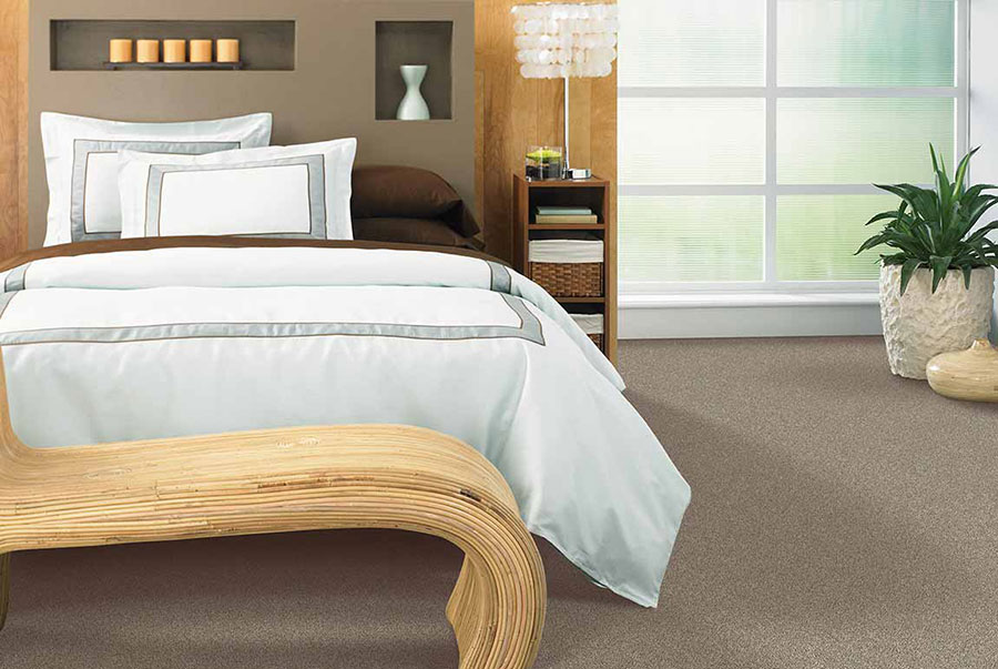 Modern themed bedroom decorated with a bed and white comforter, a modern wood ottoman, a lamp with a pendant lampshade on a nightstand, and a small potted plant on earth-toned carpet. 